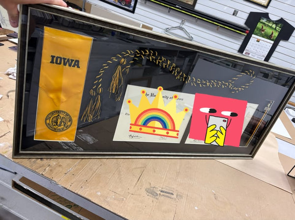 Shadow box framing an Iowa Hawkeyes pennant, a graduation tassel and children's artwork