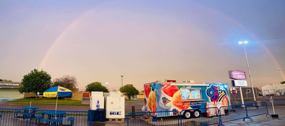 SnoBiz trailer parked outside Mahaska Drug at dusk under a rainbow